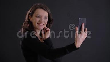 年轻迷人的高加索长发女孩在黑色背景下使用智能手机制作自拍照片的肖像。