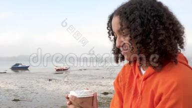 穿着橙色连帽衫的非裔美国少女少女，在港口边喝着外卖咖啡，看上去很开心