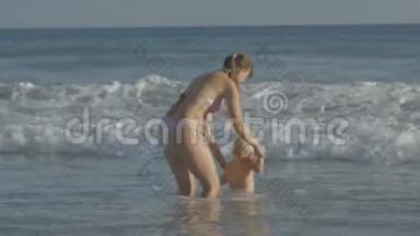 年轻的白种人母亲和两岁的金发婴儿一起玩耍，在海浪上跳跃，手牵着手在海边。 快乐