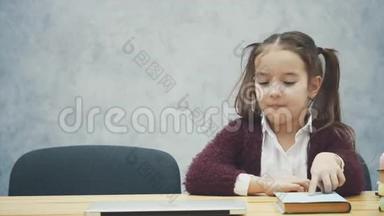 一个女学生坐在桌子旁。 在此期间，选择一本书或笔记本电脑。 在灰色背景下快乐。 开启