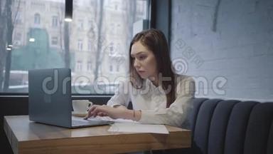 专心致志的女人用她的笔记本<strong>电脑</strong>坐在咖啡馆的桌子上。 女孩坐在靠近窗户的沙发上，<strong>端</strong>着一杯