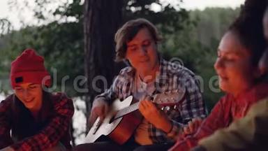 年轻英俊的游客一边弹吉他，一边微笑，他的朋友们一边唱歌，一边尽情地休息