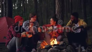 快乐的男人和女人在森林里徒步旅行时，围坐在火堆旁，煮棉花糖，弹吉他，唱歌