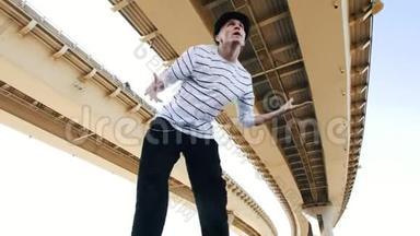 一位年轻的波普戴着帽子，穿着一件条纹T恤，在夏天在桥下跳舞，展示自由泳