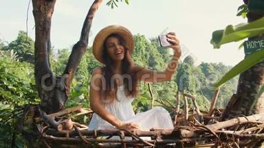年轻的微笑混合种族旅游女孩穿着白色连衣裙制作自拍照片使用手机坐在装饰草巢。