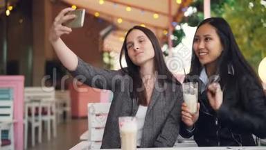 迷人的女孩正和她的亚洲朋友一起自拍，她坐在露天咖啡馆里，拿着智能手机和饮料