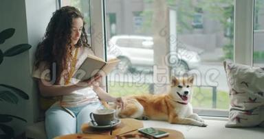 漂亮的女孩看书，抚摸小狗，在咖啡馆的窗台上放松