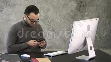 有胡子的人，他坐在一张黑色的桌子旁，看着他的智能手机，前面有他的电脑。