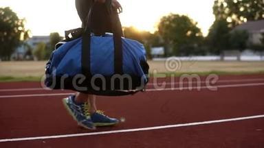 年轻的肌肉男，穿着黑色衬衫，手里拿着蓝色的运动袋，在体育场的<strong>跑道上</strong>行走。 准备接受训练