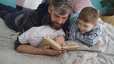 可爱的父母留着胡子的男人正<strong>在</strong>给他好奇的小<strong>儿子在家里</strong>的双人床上读有趣的书。 为人父母，快乐