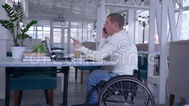 坐在轮椅上的残疾人用手机说话，拿着一台<strong>电脑</strong>工作，坐在桌子上，<strong>端</strong>着一杯