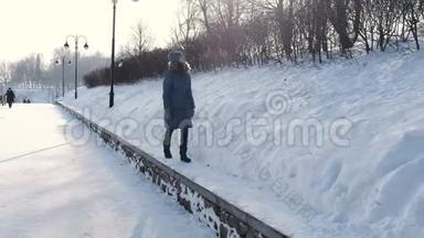 穿着蓝色<strong>羽绒服</strong>的年轻女子在冬季公园沿着边境散步，看着天空，前景。
