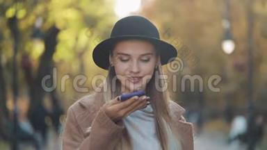 年轻漂亮的女士穿着时髦的外套，戴着黑色帽子，在秋天的公园里用智能<strong>手机</strong>发送<strong>语音</strong>信息