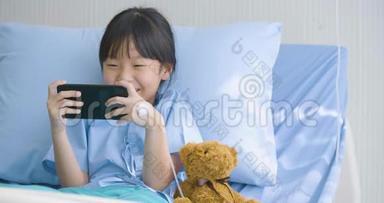 可爱的小女孩躺在医院的床上，看有趣的卡通片，智能手机上的电影。 疾病和治疗