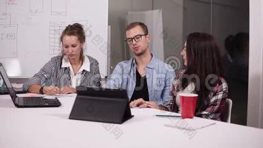 戴眼镜的年轻人和两个随意坐在办公桌前的女人，桌子上放着笔记本电脑和平板电脑，创意十足