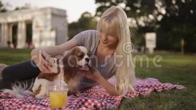 一位金发碧眼的女人躺在公园的格子布上，试图用她的猎犬拍一张自拍照片