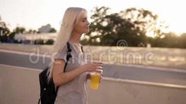 一个美丽的长发女人走在街上。 带着黑色背包和一杯柠檬水的放松女孩