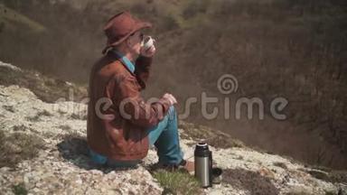 山上坐着一个男人，戴着牛仔帽、皮夹克、蓝色牛仔裤和眼镜。 一个人<strong>喝热水</strong>瓶里的茶。