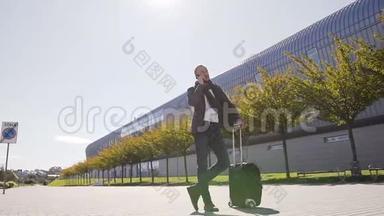 年轻的<strong>时尚男士</strong>在离开机场时拿着手提箱站在手机上说话。 户外。 商业<strong>风</strong>格