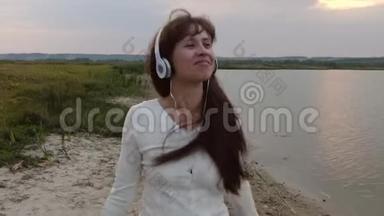 戴着耳机的快乐女孩听音乐和跳舞。 美丽的女孩晚上在沙滩上玩平板电脑