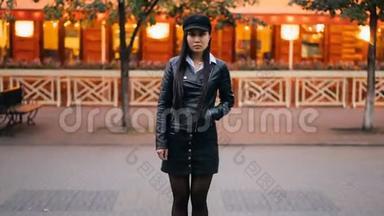 一个漂亮的亚洲女孩看着摄像机站在户外，手插口袋，穿着时髦的皮革