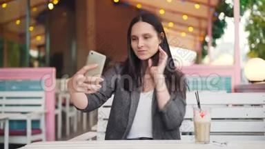 一位留着深色长发的漂亮女孩正在一个人坐在<strong>街头</strong>咖啡馆里自拍并为智能手机<strong>拍照</strong>