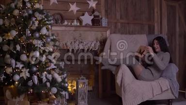 在圣诞节那天，女孩拿着礼物打开盒子，坐在圣诞树附近的椅子上高兴。 4K