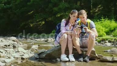 年轻的游客看着相机上拍摄的照片。 他们坐在靠近山河和森林的岩石上