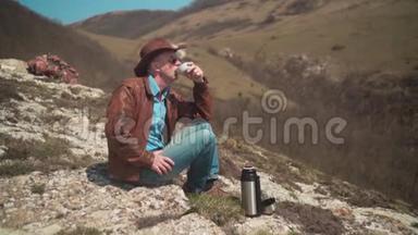 山上坐着一个男人，戴着牛仔帽、皮夹克、蓝色牛仔裤和眼镜。 一个人喝热水瓶里的茶。