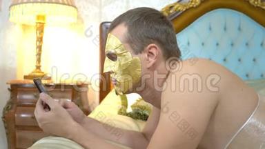 戴着<strong>金色</strong>面罩的英俊男子正在使用<strong>手机</strong>在豪华的床上放松。 特写