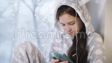 女孩少年冬天穿着温暖的睡衣坐在<strong>窗边</strong>，用你的智能手机给朋友发短信。 <strong>窗</strong>外