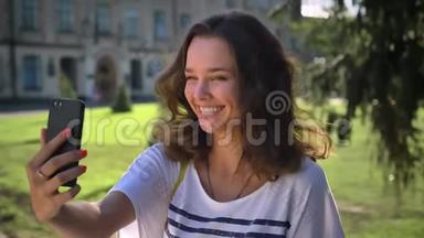 一位年轻微笑的白种人女孩站在公园里，用智能手机打招呼视频聊天，制作的肖像。