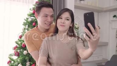 亚洲夫妇使用智能手机自拍与圣诞树装饰他们的客厅在家里圣诞节。