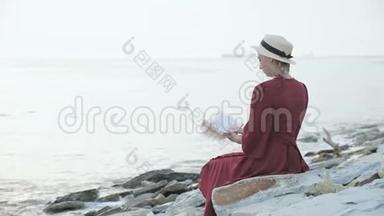 日落时分，一个漂亮的白人女孩穿着红色鲜艳的裙子和一顶草帽坐在海边的一块大石头上