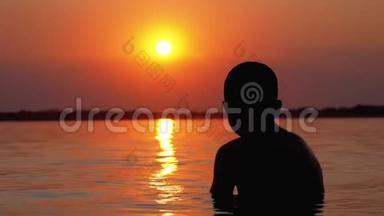 日落和橙径背景下坐在水里的男孩剪影。 慢动作