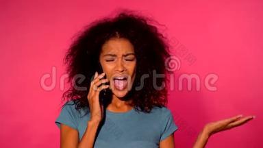 愤怒的非洲裔美国妇女，一头卷发，穿着绿色t恤，在粉色墙背景下用智能手机打电话