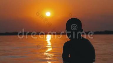 日落和橙径背景下坐在水里的男孩剪影。 慢动作