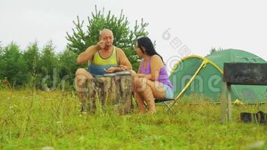 一个男人和一个女人在帐篷附近坐在一个木头树桩旁边，在雨天从盘子里吃东西。