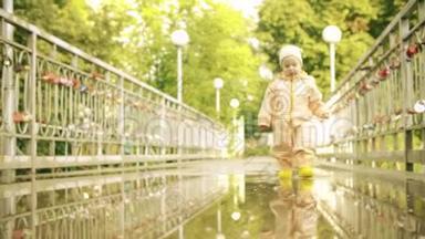 穿着橙色防水雨衣和橡胶雨靴的滑稽小女孩在雨后的水坑里慢动作