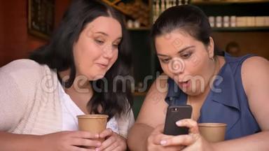 两个多肉的白种人女孩在咖啡馆里拿着纸杯，在感情上讨论着看电话