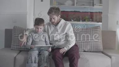 父亲和孩子坐在大客厅的沙发上，在平板电脑<strong>屏幕</strong>上<strong>滚动</strong>。 爸爸教他的孩子。 父子关系