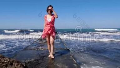 年轻的微笑快乐的女人赤脚走在海上码头上。 红色连衣裙随风飘扬。 波浪撞击海港。 慢动作