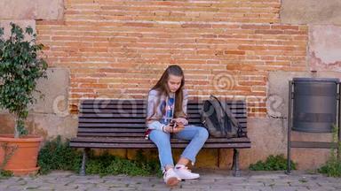 迷人的年轻女孩独自坐在栏杆上，带着手机和背包。 无聊少年滚动，发送信息