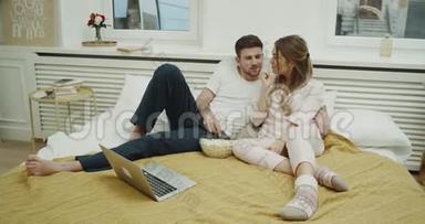 现代公寓里的年轻夫妇在卧室里<strong>一起</strong>玩，在床上吃爆米花，在笔记本电脑上<strong>看电影</strong>。