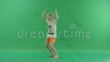 一个穿休闲服装的金发小男孩正在听音乐和跳舞。 他侧着身子站在绿色屏幕上