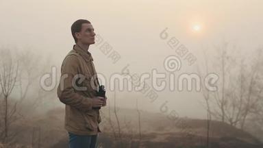 一个身穿<strong>卡其色</strong>外套的年轻红发男子，手持望远镜，看日出和日落。 四周都是雾