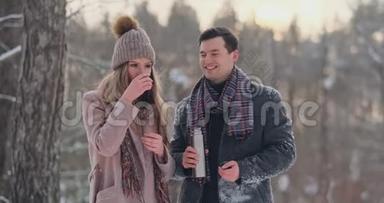 穿着外套和围巾的漂亮时髦的夫妇在冬天的森林里喝热水瓶里的茶