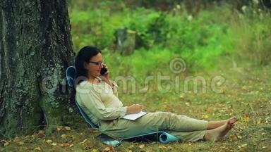 戴着眼镜的微笑着的女人光着脚<strong>坐在</strong>公园的一棵<strong>树下</strong>，用铅笔在笔记本上画画，还在说话