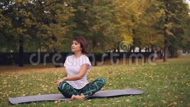 柔柔女瑜伽尼正在练习简单的扭转姿势，然后坐在公园草地上的瑜伽垫上以莲花的姿势休息