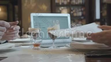 无法辨认的男人和女人坐在咖啡馆的桌子旁工作。 笔记本电脑、文件和带咖啡的眼镜都在上面